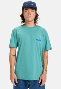 Camiseta de algodón turquesa con mangas cortas y cuello redondo, con un logotipo gráfico azul en la parte izquierda del pecho. Lleva puesta un pantalón de mezclilla azul.