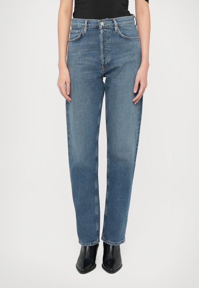 Aukštakulnės mėlynos džinsinės kelnės su tiesiomis kojų linijomis. Klasikinio penkių kišenių dizaino ir metalinės sagos užsegimas. Dėvimos su juodais batais.