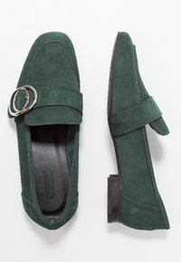 Ballerines en suédine verte avec un bout rond, accent décoratif en anneau argenté et un petit talon en caoutchouc noir. Des coutures contrastées sont présentes.