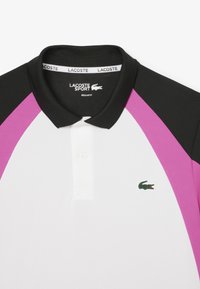 Polo shirt avec un col et des manches noires, un corps blanc et des panneaux d'épaule roses. Logo de crocodile vert brodé sur la poitrine. Conception à coupe régulière.