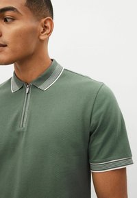 Grøn polo t-shirt i tekstureret stof, med to-knaps lynlås krave og stribede detaljer ved kraven og ærmerne. Nærbillede viser syningerne.
