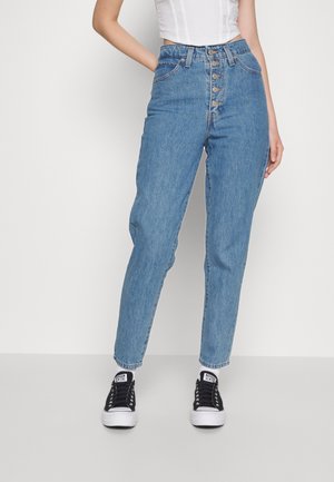 Vrouw draagt een blauw spijkerbroek met hoge taille en knoopsluiting, zwart-witte sneakers en een wit mouwloos topje, staande tegen een witte achtergrond.