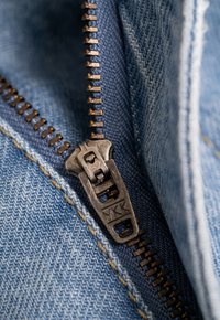 Cremallera de metal en tela de denim, con un tirador de la marca YKK, en tonos azules y costuras visibles a lo largo de los bordes.