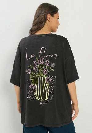 Kobieta stojąca tyłem, ubrana w czarny, oversize'owy T-shirt z różowym i zielonym wzorem kwiatów w wazonie oraz tekstem "Les Fleurs", "Flower Market", "1987", "Paryż, Francja".