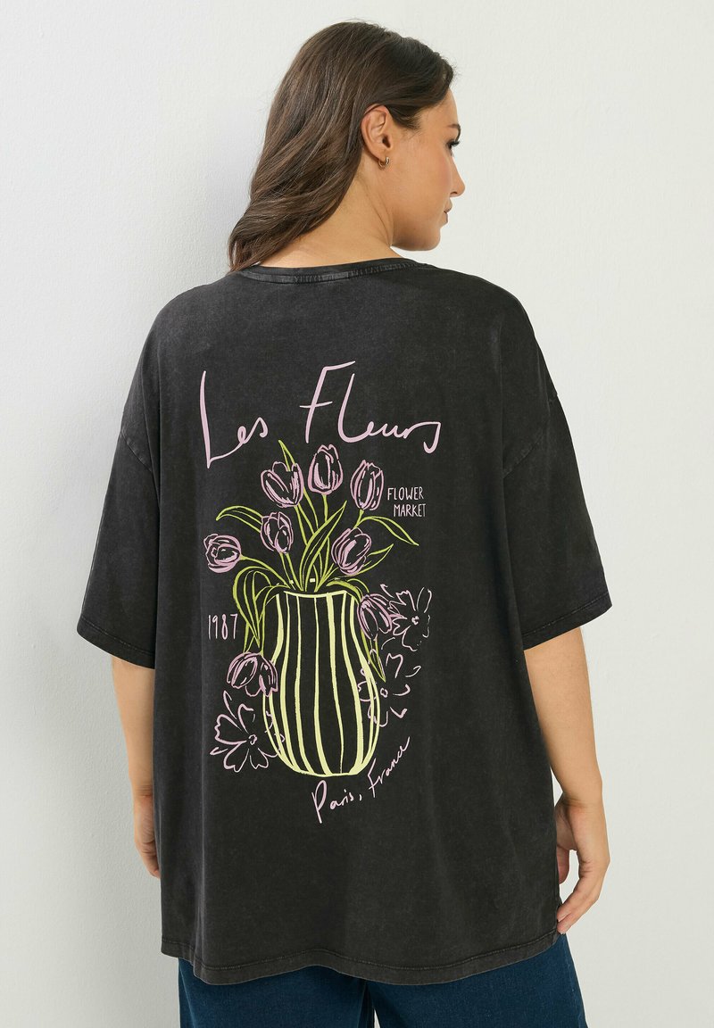 Kobieta stojąca tyłem, ubrana w czarny, oversize'owy T-shirt z różowym i zielonym wzorem kwiatów w wazonie oraz tekstem "Les Fleurs", "Flower Market", "1987", "Paryż, Francja".