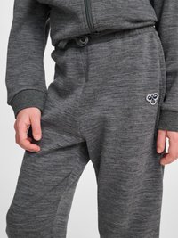 Barn iført grå joggingbukser og matchende lynlåstrøje, hænderne afslappet ved siderne, bi-logo synligt på venstre lår.