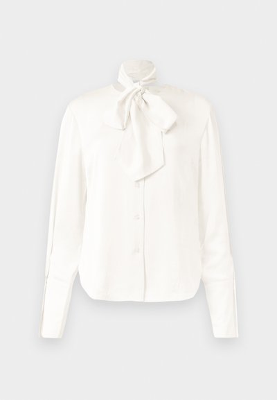 Blouse blanche à manches longues en tissu léger avec col haut et un grand nœud au niveau du cou, dotée d'une fermeture à boutons sur le devant.
