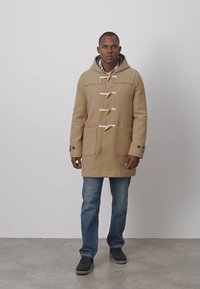 Beige duffelcoat i ullblandning med träknappar, stora fickor och fodrad huva, bärs över blå jeans och svarta skor.