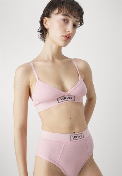 Lys rosa ribbet bralette og højhalsede trusser, med en sort Versace-logo-bånd, tynde stropper og en blød tekstur.