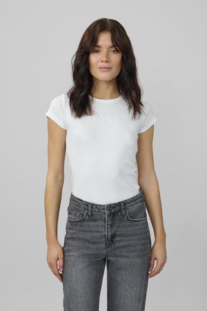 Jonge vrouw met schouderlang golvend bruin haar, gekleed in een wit nauwsluitend T-shirt met korte mouwen en een hoog uitgesneden grijze spijkerbroek, staand tegen een grijze achtergrond.