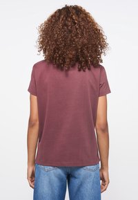 Persona con cabello rizado que lleva una camisa de manga corta color burdeos y jeans azules, vista desde atrás contra un fondo blanco liso.