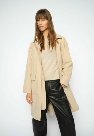 Kvinde med langt brunt hår iført en beige frakke, lys sweater og sorte læderbukser, stående mod en ensfarvet lys baggrund.