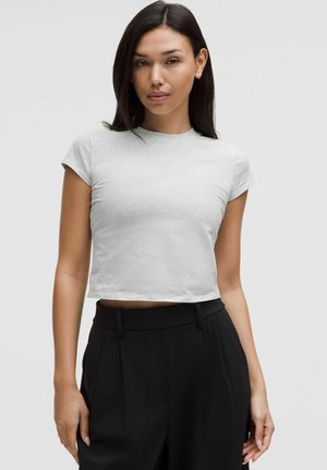 Kvinde med langt, mørkt hår iført en tætsiddende, lysegrå crop top med korte ærmer og højttaljede sorte plisserede bukser mod en ensfarvet baggrund.