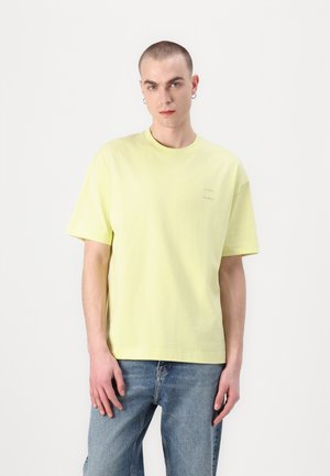 Camiseta de color amarillo claro, de manga corta, con un cuello redondo y un sutil logo. Combinada con jeans azules, de pie frente a un fondo blanco.