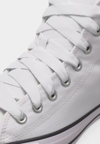 Κοντινό πλάνο λευκού καμβαδένιου αθλητικού παπουτσιού που δείχνει υφή υφάσματος, λευκά κορδόνια περασμένα μέσα από ασημένιες τρύπες και μέρος του λάστιχου της σόλας.