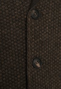 Brązowa tkanina o fakturze z wzorem plecionym, z dwoma czarnymi guzikami i płaskim kołnierzem. Zbliżenie na przedni detal ubrania.