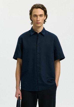 Ung mann som har på seg en mørkeblå kortermet skjorte med knapper og svarte bukser, står med den ene hånden i lomma og holder en mørk gjenstand.