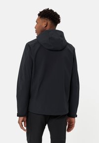 Black softshell jacket with a hood, featuring a smooth texture, rear seam details, and elastic cuffs, worn by a person from behind.