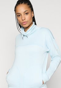 Svetlo modrý pulóver s vysokým golierom, s rebrovanou textúrou, prednými vreckami a logom na rukáve. Vyrobený z mäkkého materiálu.