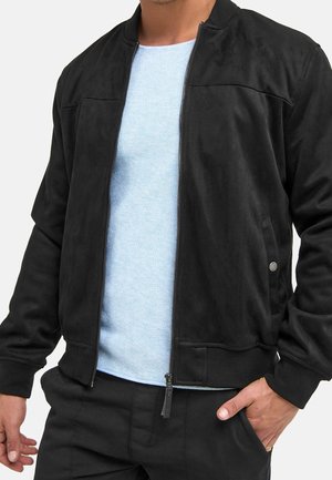 Mann trägt schwarze Reißverschlussjacke über hellblauem strukturiertem Hemd, eine Hand in der Tasche vor neutralem Hintergrund.