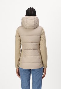 Beige prošívaná bunda s velkou kapucí, se zaoblenými prošívanými sekcemi na zádech a fitted rukávy, doplněná modrými džíny.