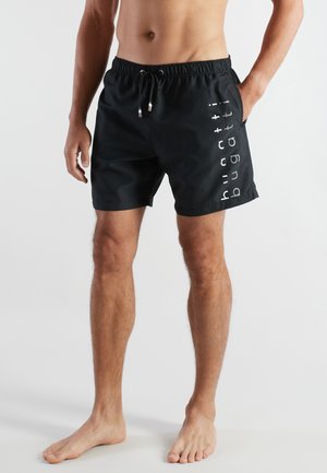 Mann trägt schwarze Badeshorts mit senkrechtem "Billett"-Logo am linken Bein, steht barfuß vor einem schlichten Hintergrund.