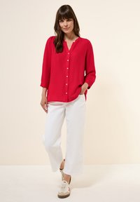Blusa rossa con bottoni, scollatura a V e maniche a tre quarti. Abbinata a pantaloni bianchi a gamba larga e sneakers beige.