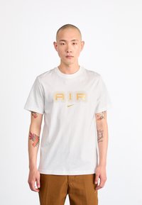 Bijela pamučna majica s natpisom "AIR" u zlatnim slovima i malim Nike logom. Okrugli izrez, kratki rukavi i opušten fit.