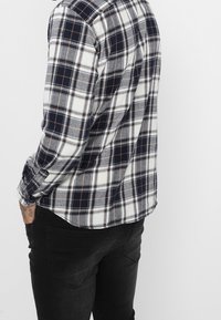 Koszula na guziki w kratę w odcieniach granatu, bieli i szarości, wykonana z miękkiej tkaniny, z mankietami zapinanymi na guziki i zaokrąglonym suwakiem. Noszona z ciemnymi dżinsami.