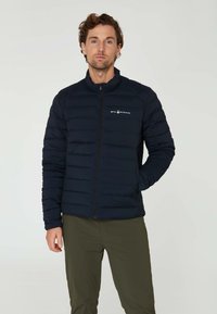 Marinblå pufferjacka med dragkedja, horisontellt quiltad design och hög krage. Har en liten logotyp på vänster bröst. Bärs med olivgröna byxor.