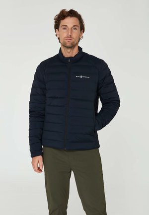 Doudoune navale avec fermeture éclair, design matelassé horizontal et col haut. Présente un petit logo sur la poitrine gauche. Portée avec un pantalon olive.