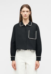 Mujer con camisa negra corta con ribete de perlas blancas en el cuello, bolsillo y puños, combinada con pantalones blancos de cintura alta.