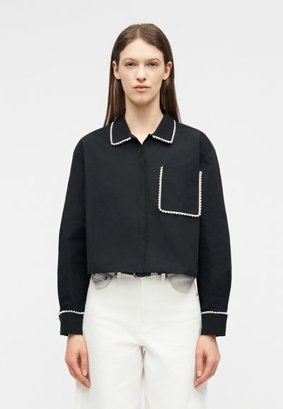 Γυναίκα που φοράει μαύρο κοντό πουκάμισο με λευκή περιμετρική διακόσμηση από μαργαριτάρια στον λαιμό, την τσέπη και τις μανσέτες, συνδυασμένο με λευκό παντελόνι ψηλής μέσης.