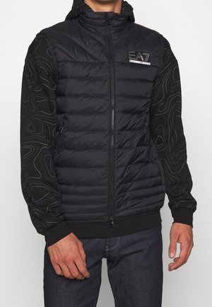 Homme portant un gilet noir matelassé zippé avec des manches à motifs et le logo EA7 sur la poitrine gauche, associé à un jean foncé, debout devant un fond blanc.