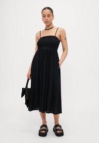 Juodas be rankovių Suknelė su susiraukšlėjusiu kūnu, laisvai krintančia sijonu ir kišenėmis. Suderinta su masyviomis juodomis šlepetėmis su dekoratyviniais metaliniais elementais ir juoda rankine.