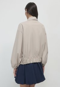 Frau mit langen dunklen Haaren, die eine beige kurzes Jacke mit elastischem Saum und Ärmeln sowie einen marineblauen Faltenrock trägt, von hinten gezeigt.