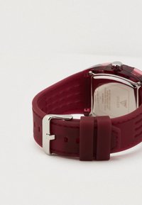 Rød silikoneremur med kvadratisk urskive, sølvfarvet metalspænde, diskret tekstur på remmen og indgraveret mærke på bagsiden af kassen.