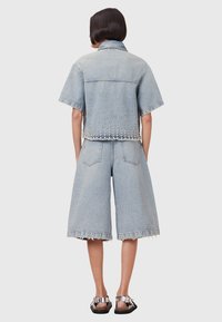 Giacca cropped in denim blu chiaro con maniche corte, con accenti simili a perle lungo l'orlo, abbinata a pantaloni corti fino al ginocchio dello stesso colore.