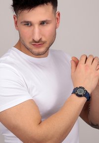 Orologio digitale con quadrante rotondo nero, cinturino in resina blu e accenti metallici; modello indossa una t-shirt bianca aderente, che mette in mostra un braccio muscoloso.