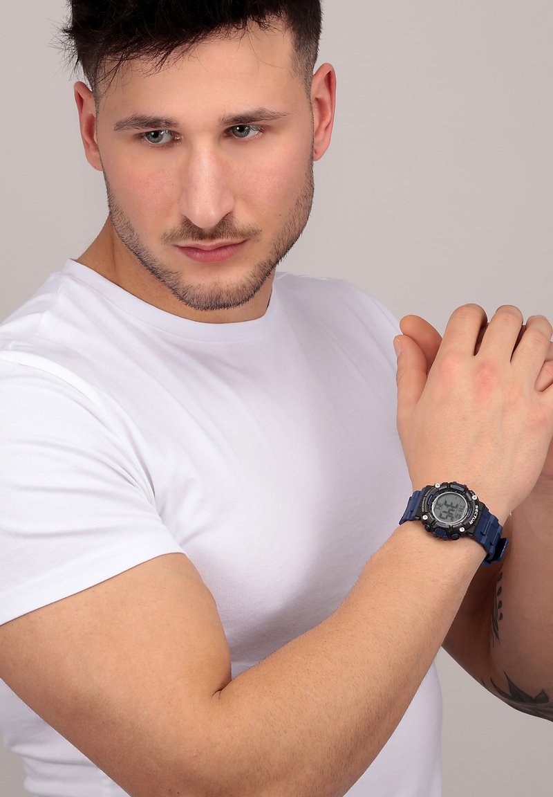 Orologio digitale con quadrante rotondo nero, cinturino in resina blu e accenti metallici; modello indossa una t-shirt bianca aderente, che mette in mostra un braccio muscoloso.