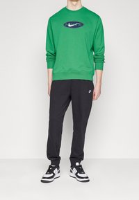 Grön Nike-tröja med ribbade muddar, kombinerad med svarta Nike-mjukisbyxor. Skorna har en vit och svart design som framhäver en sportig stil.