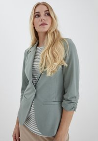 Ljusgrön blazer med en figursydd design, uppkavlade ärmar och två framfickor, kombinerad med en randig topp under.