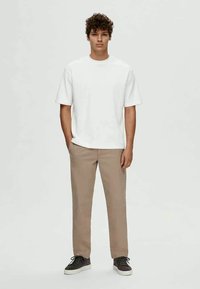 T-shirt branca de manga curta com gola redonda, combinada com calças bege claro. Os sapatos são escuros com solas brancas. Design simples e casual.