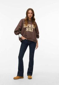 Sudadera marrón con la inscripción "APRES SKI" en crema, combinada con vaqueros acampanados de mezclilla oscura y zuecos color camel. Textura suave, ajuste relajado.