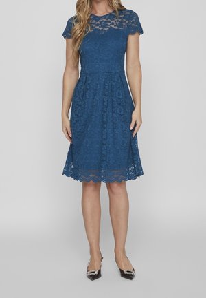 Femme portant une robe en dentelle bleue mi-longue, à manches courtes, cintrée à la taille avec un ourlet festonné, associée à des escarpins noirs pointus ornés d'embellissements.