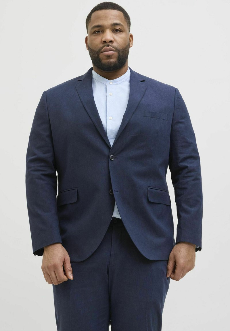 Dunkelblazer in Marineblau mit strukturierter Oberfläche, ausgestattet mit einer Brusttasche und zwei Fronttaschen, getragen über einem hellblauen Hemd mit Kragen.