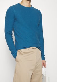 Blå tröja med lång ärm och rund hals i texturerat tyg, kombinerad med beiga byxor. Modellen håller en vit tygväska.