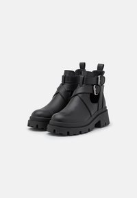 Botas negras por el tobillo hechas de cuero suave con una suela gruesa, que cuentan con dobles correas ajustables con hebillas metálicas y detalles laterales recortados.