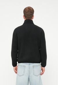 Uomo che indossa un giubbotto in pile nero con colletto alto e jeans azzurri chiari, in piedi con le spalle rivolte verso la fotocamera su uno sfondo semplice.