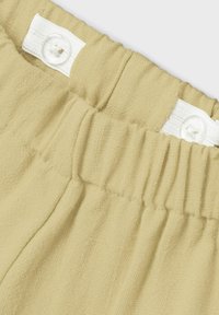 Cintura elastica beige chiaro con texture arricciata e chiusura a bottone bianco. Il tessuto appare morbido e leggermente texturizzato.